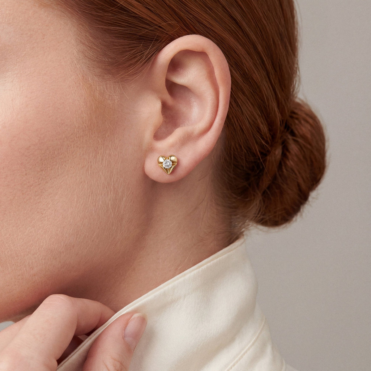 Woman in an ivory silk shirt with a turned-up collar wearing a natural diamond heart stud earring in 14K yellow gold