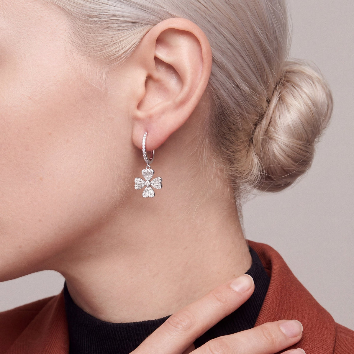 Woman in a rust blazer over a black high neck wearing a natural diamond clover drop huggie earring in 18K white gold, hand on the lapel