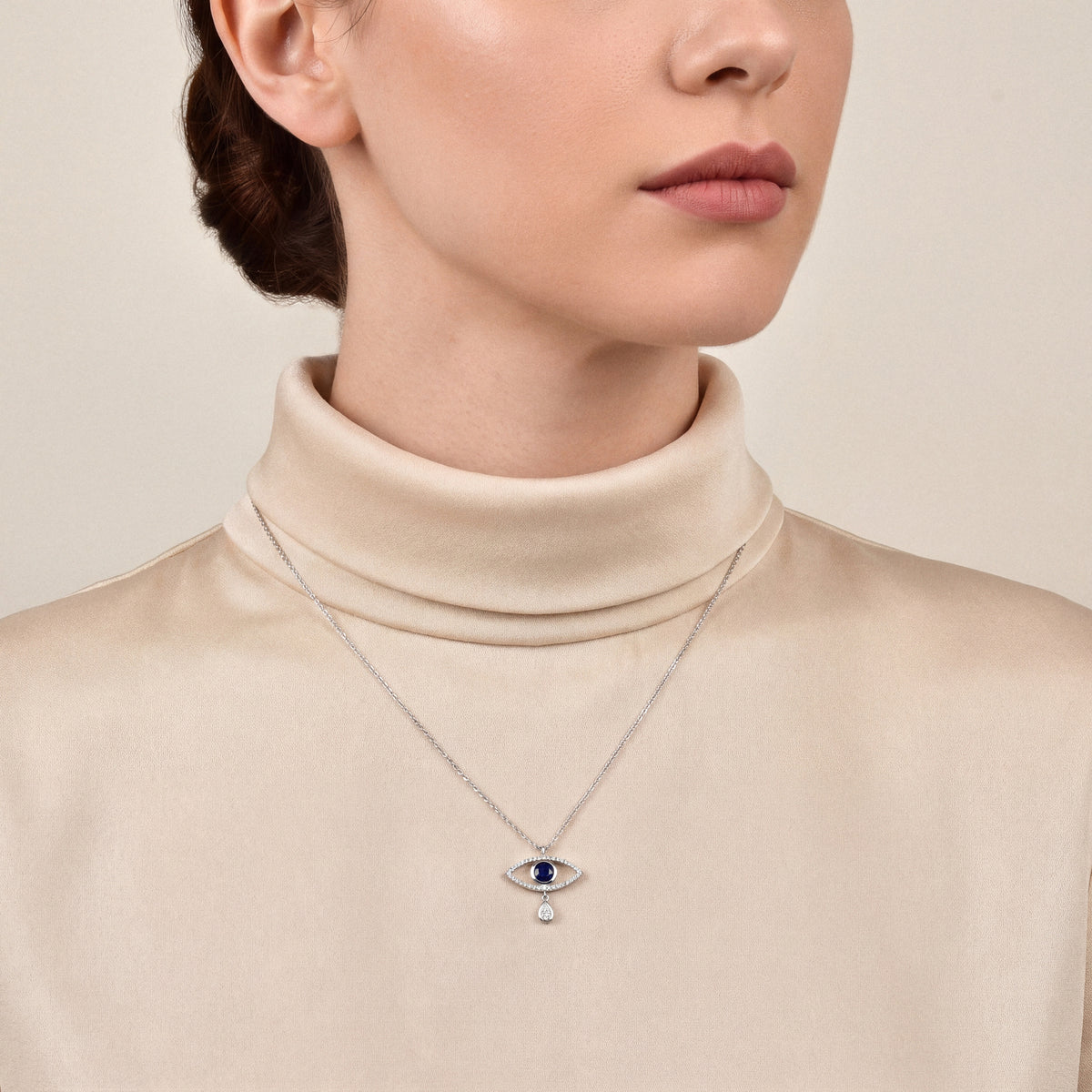 Woman in a champagne silk funnel-neck top wearing an evil eye blue sapphire and natural diamond pendant necklace in 14K white gold