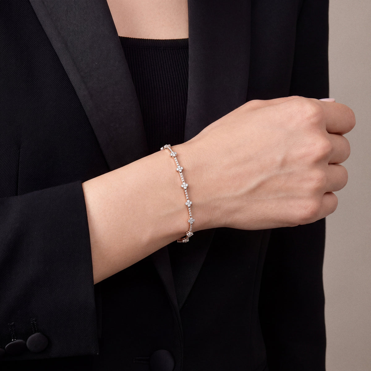 Woman in a black tuxedo jacket wearing a natural diamond flower station bracelet in 14K rose and white gold, loose fist at the waist
