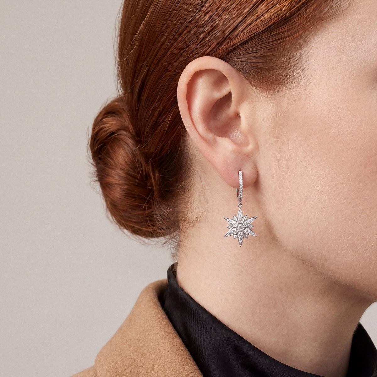 Woman in a camel blazer over a black mock neck wearing a star natural diamond drop huggie earring in 14K white gold