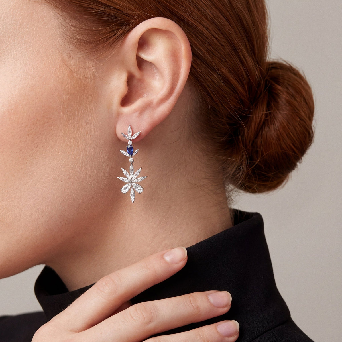 Model in a black blazer and high collar in profile, fingertips at her collar, wearing a natural diamond and sapphire flower drop earring in 14K white gold