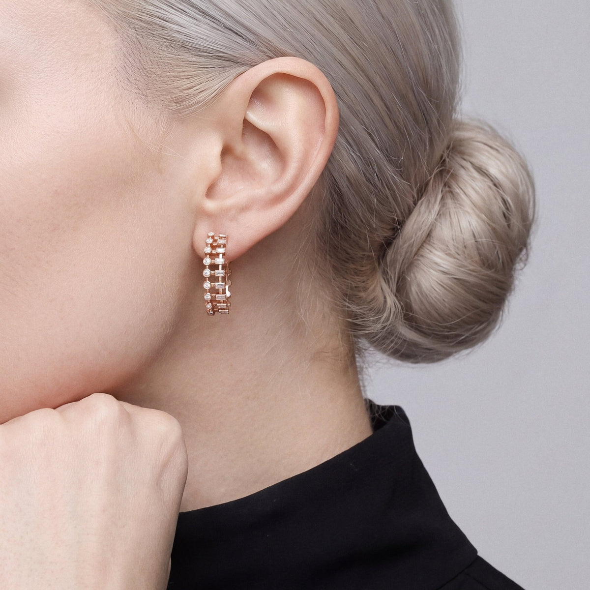 Model in a black high-neck top resting her fist under her jaw, wearing a baguette and round natural diamond hoop earring in 14K rose gold