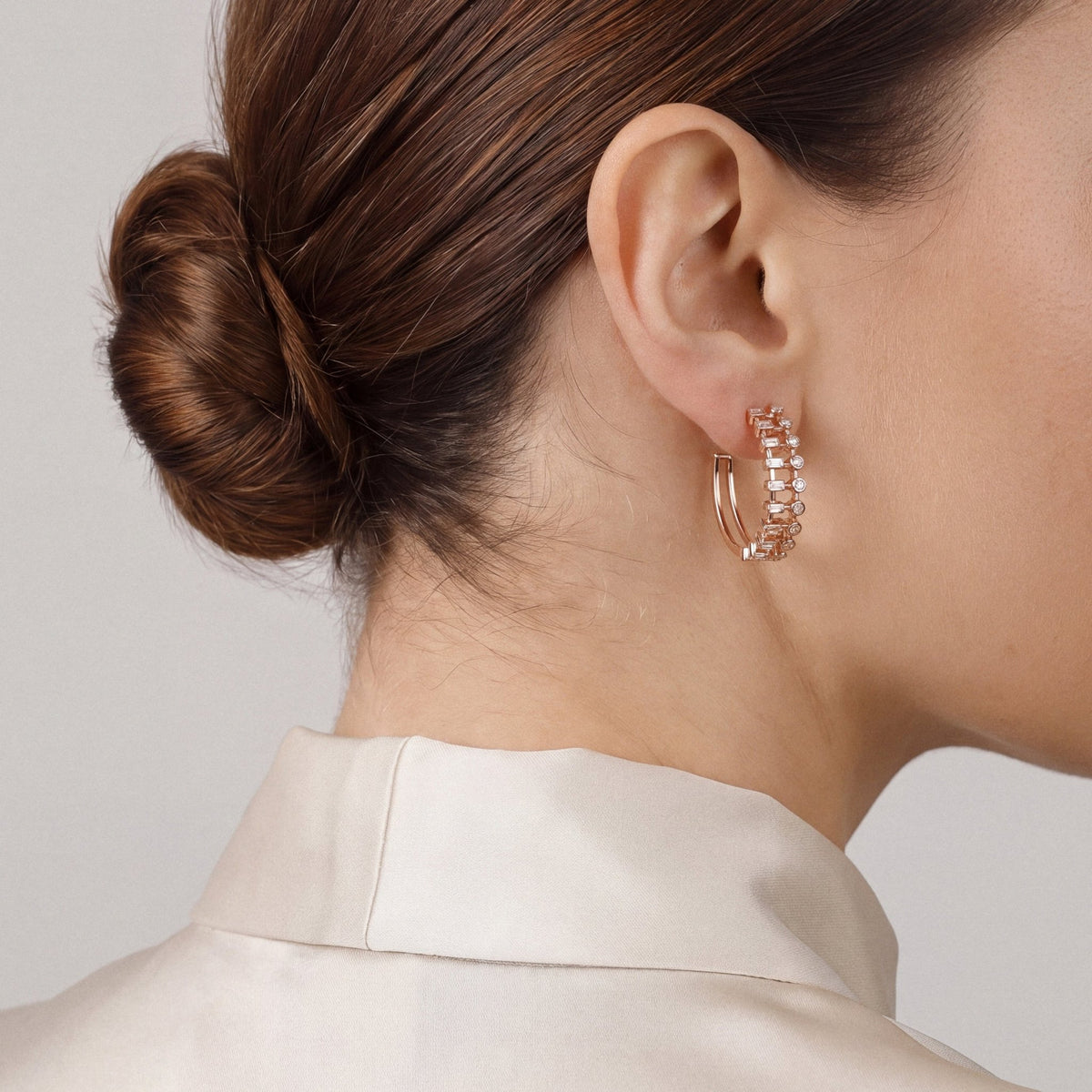Model in an ivory satin stand-collar blouse, turned away, wearing a baguette and round natural diamond hoop earring in 14K rose gold