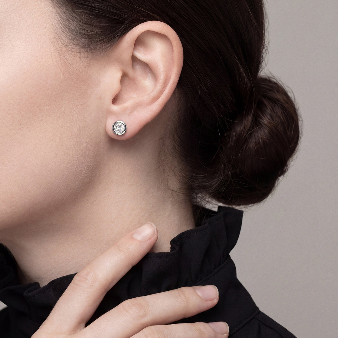 Woman in a black ruffle-collar blouse, fingertips at her collar, wearing a bezel-set round natural diamond solitaire stud earring in 14K white gold