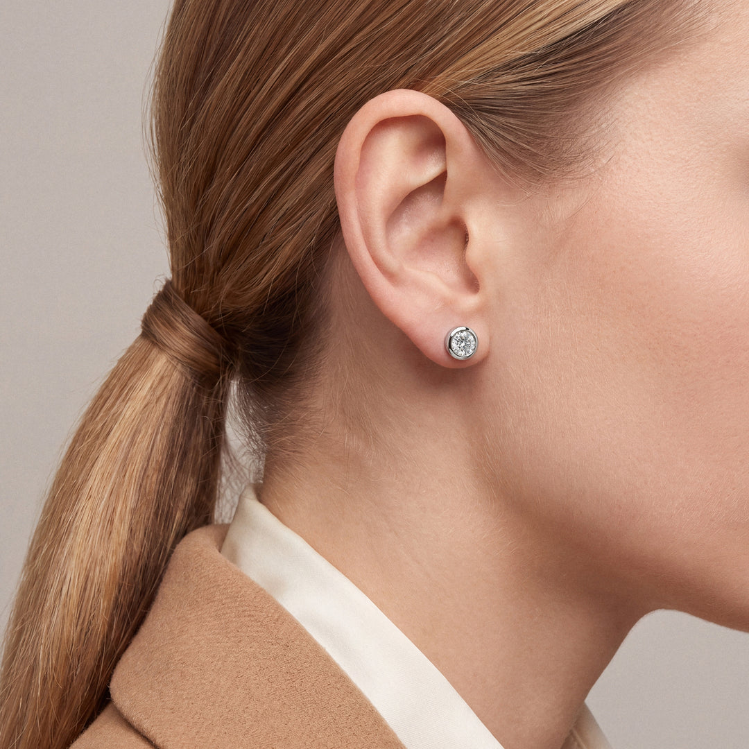 Woman in profile in a camel blazer over an ivory shirt wearing a bezel-set round natural diamond solitaire stud earring in 14K white gold