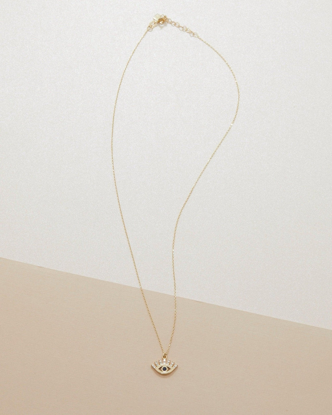 The 14K solid gold evil eye necklace laid out at full length on a white and beige background.