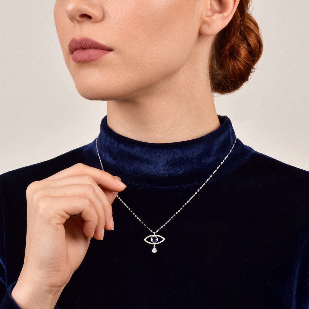 Woman in a midnight-navy velvet mock-neck top holding the chain of an evil eye blue sapphire and natural diamond pendant necklace in 14K white gold