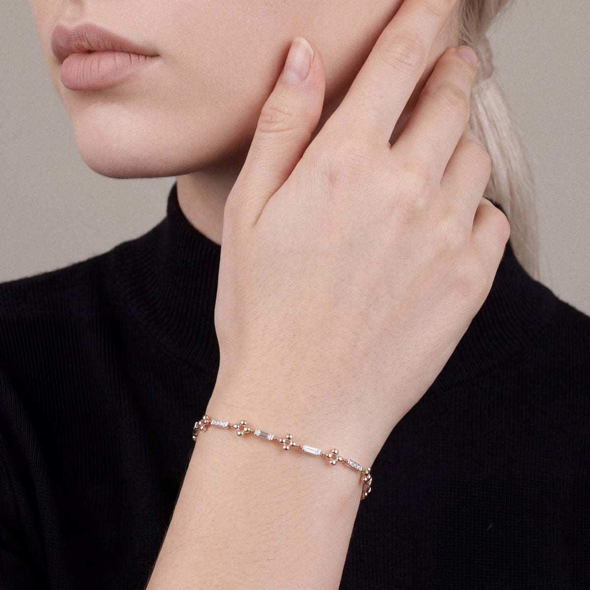 Woman in a black mock-neck knit wearing a natural diamond baguette flower station bracelet in 14K white and rose gold, hand at the cheek