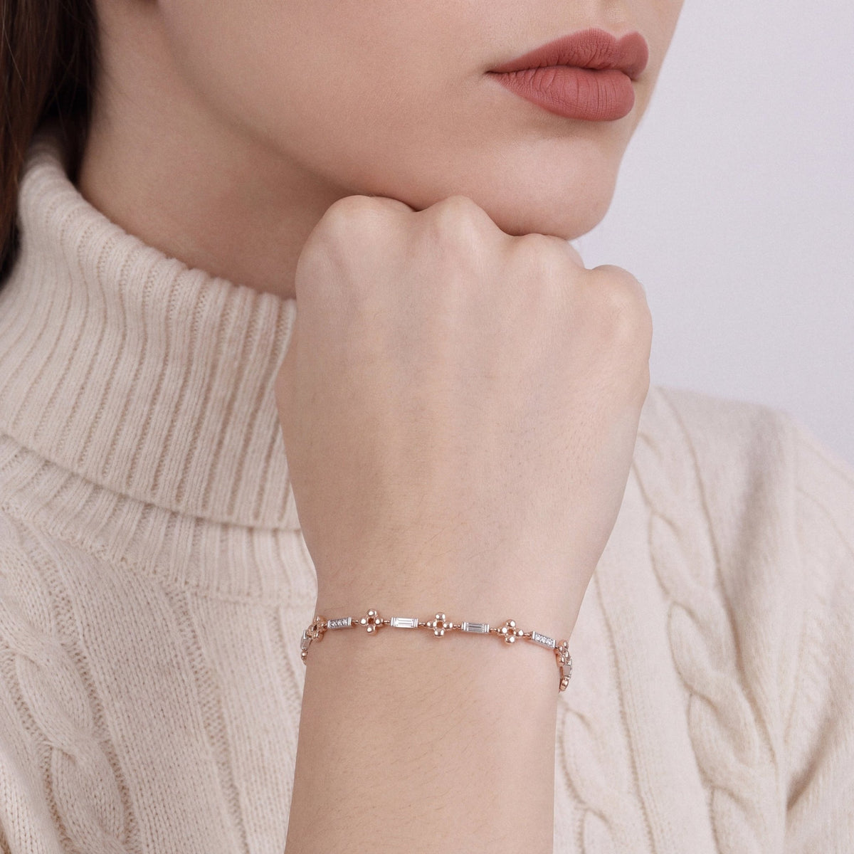 Woman in a cream cable-knit turtleneck wearing a natural diamond baguette flower station bracelet in 14K white and rose gold, fist resting under the chin