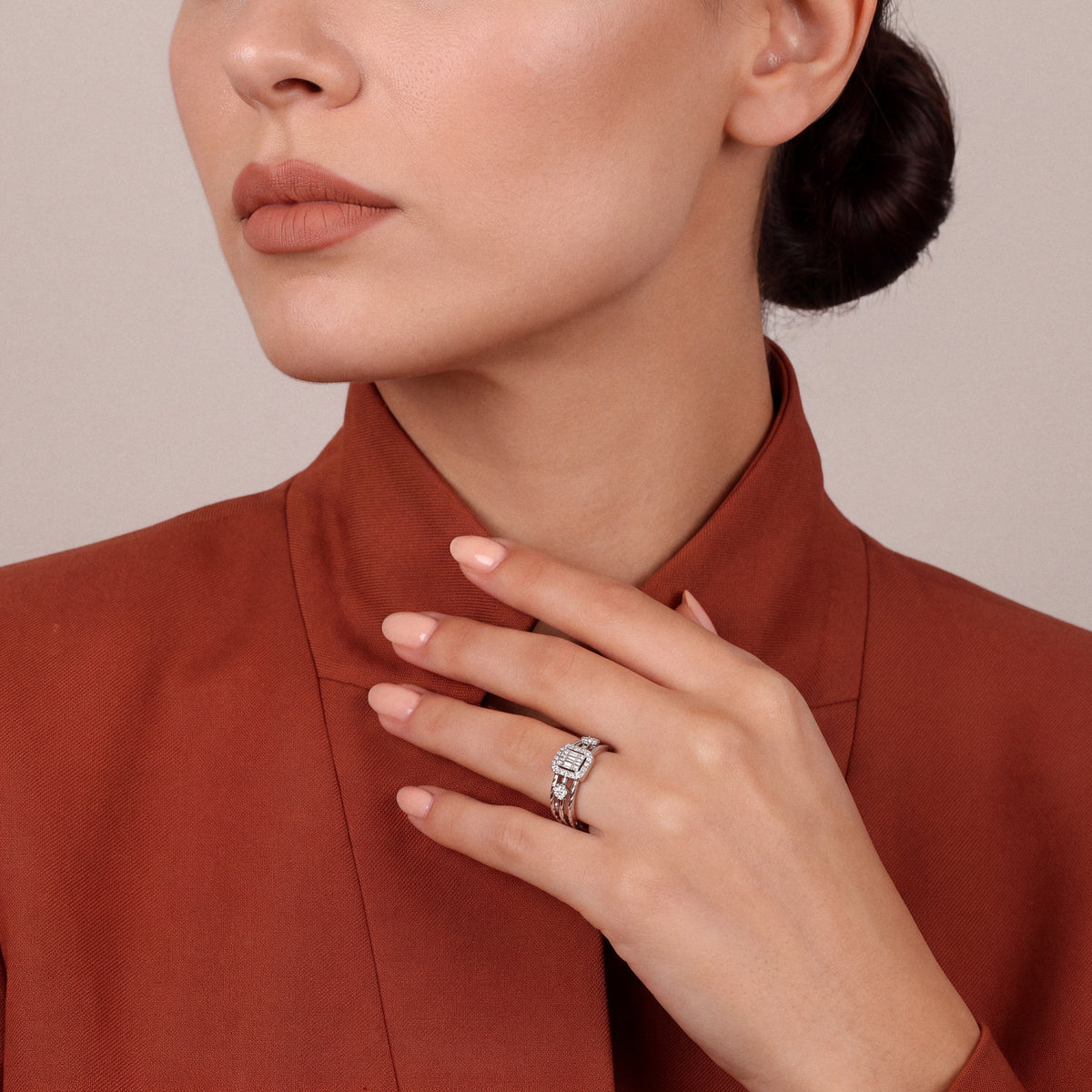 Woman in a rust stand-collar jacket wearing a natural diamond baguette halo ring, fingertips at the collar