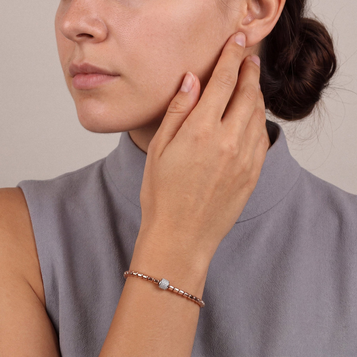 Woman in a dove-grey sleeveless funnel-neck top, hand at her cheek, wearing the natural diamond pavé barrel bead bracelet in 14K rose and white gold