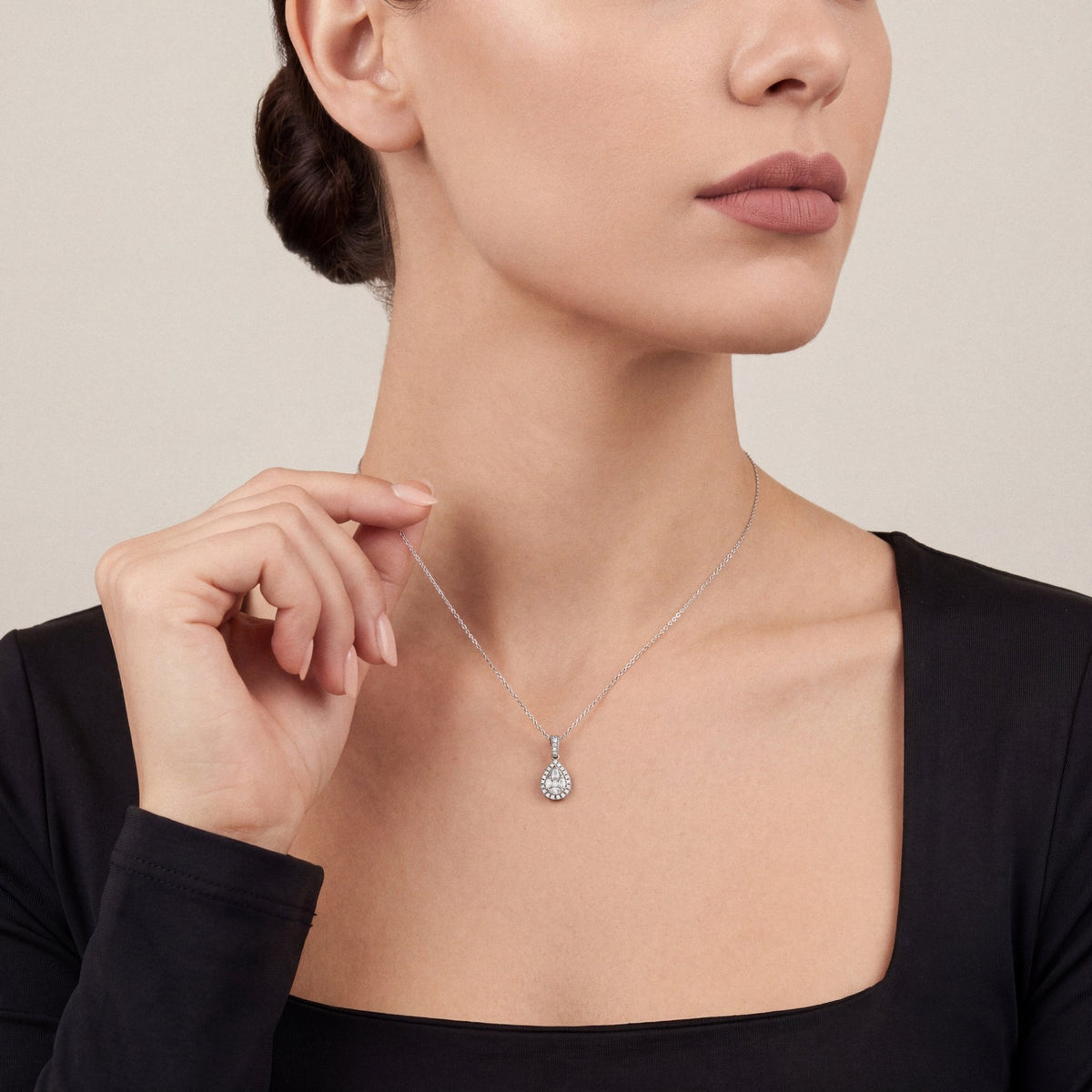 Model in a black crepe square-neck top lifting the chain of a 0.51 ct natural diamond pear halo pendant necklace in 14K white gold