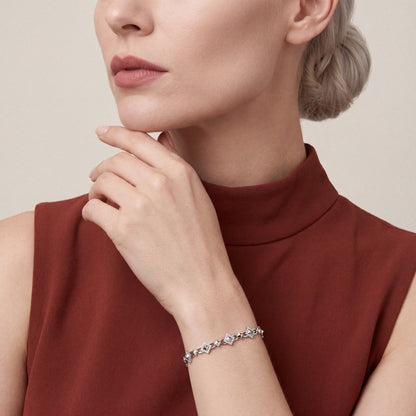 Woman in a rust sleeveless mock-neck top, fingertips at her chin, wearing a 0.92 ct natural diamond pyramid station link bracelet in 14K white gold
