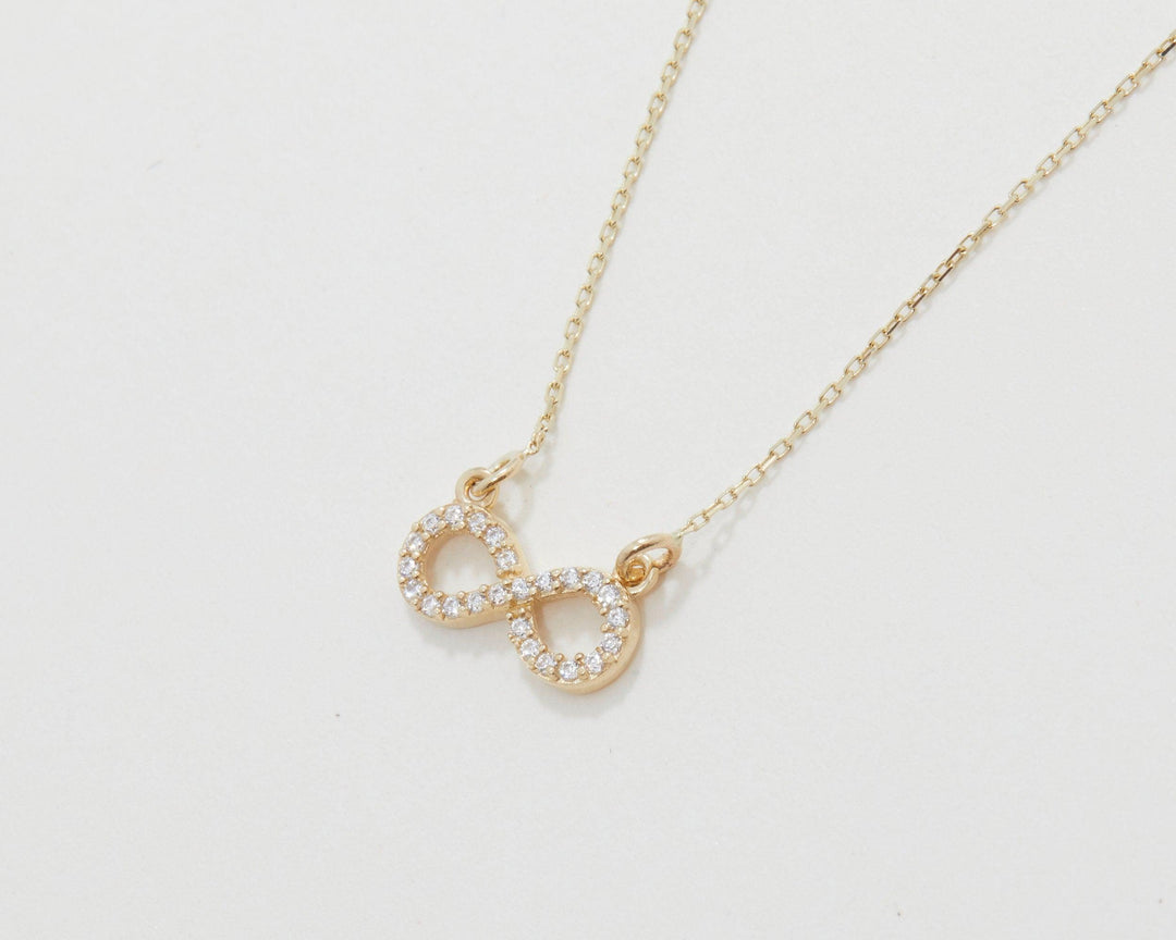 The 14K solid gold pavé infinity pendant and its chain lying flat on a light background.