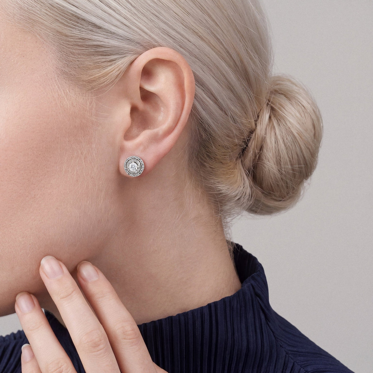 Woman in a navy pleated high-neck top, fingertips at her jaw, wearing a round natural diamond double halo stud earring in 14K white gold