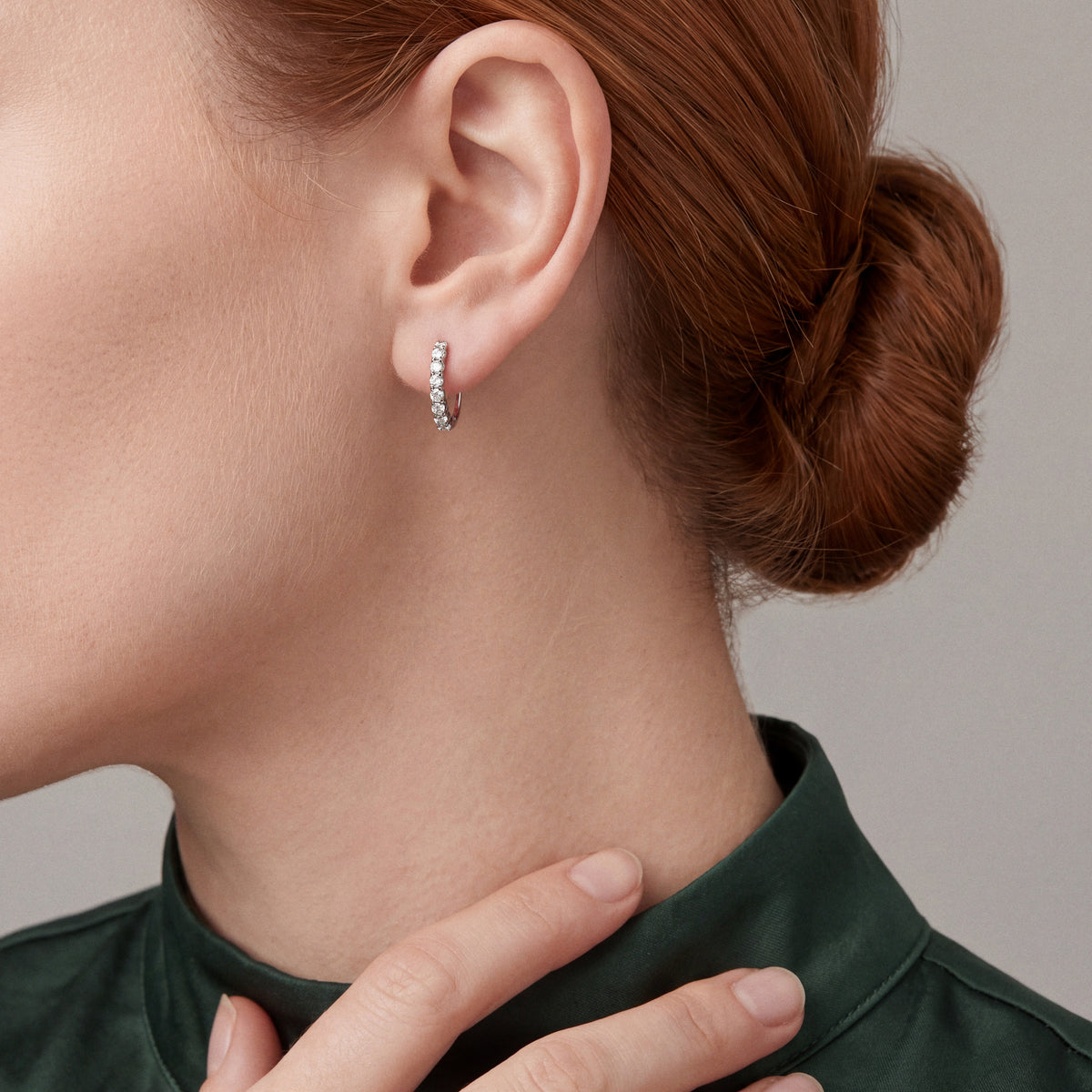 Woman with her fingertips on the collar of a forest-green silk shirt wearing a round natural diamond huggie hoop earring in 14K white gold