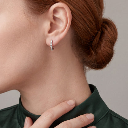 Woman with her fingertips on the collar of a forest-green silk shirt wearing a round natural diamond huggie hoop earring in 14K white gold
