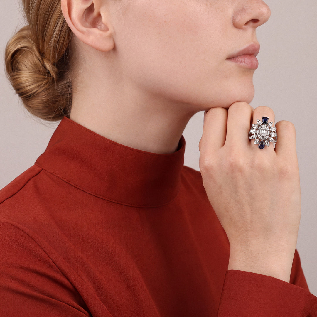 Sapphire and natural diamond baguette cocktail ring in 14K white gold worn on the ring finger, loose fist under the chin, rust silk high-collar shirt