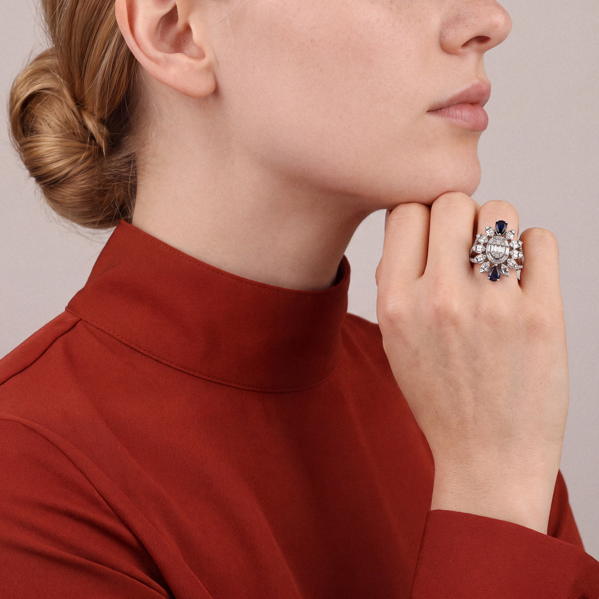 Sapphire and natural diamond baguette cocktail ring in 14K white gold worn on the ring finger, loose fist under the chin, rust silk high-collar shirt